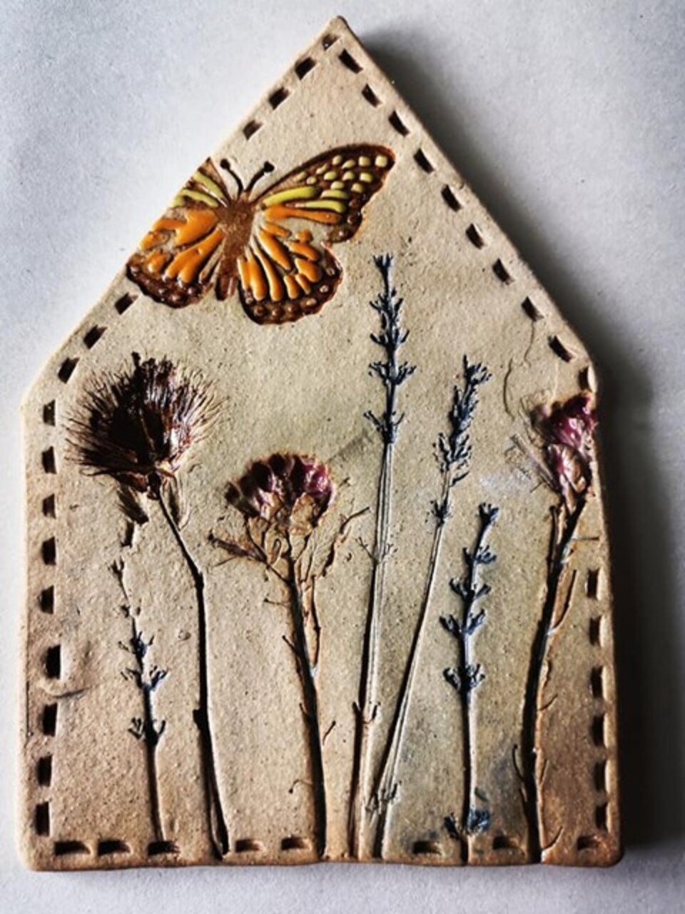 A handcrafted clay plaque shaped like a house, featuring an embossed orange and yellow butterfly at the top. Below the butterfly, several dried flowers and plants, including thistles and lavender, are embedded into the clay.